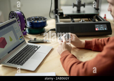 Hands of young male engineer or designer holding geometric figure over wooden table while sitting in front of laptop Stock Photo