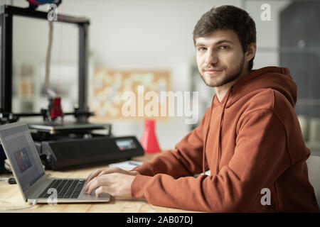 Bearded guy in brown hoodie sitting in front of laptop in office while working over sketches of items for 3d prirnting Stock Photo