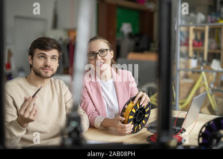 Two young creative designers looking at 3d printer while discussing its characteristics and model by workplace Stock Photo