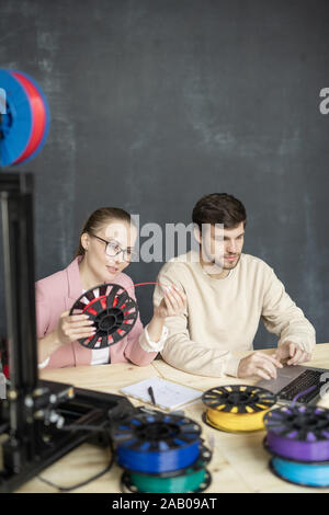 Young female looking at red filament on spool while sitting next to her colleague surfing for websites in front of laptop Stock Photo