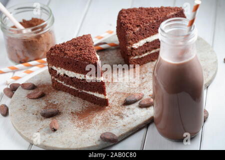 Two slices of chocolate cake, hot chocolate in a bottle, powdered cocoa and cocoa beans on a rustic table Stock Photo