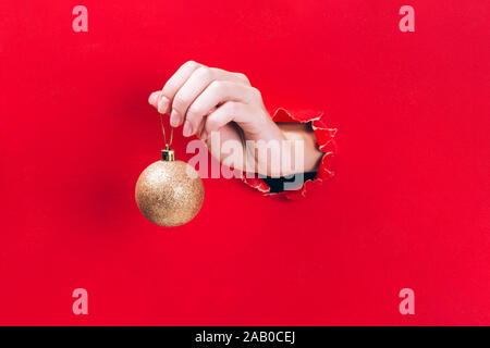 Female hand holding golden Christmas tree bauble through a hole in red background. Stock Photo