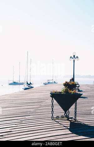 Wood pier with triangular pot, street lamps and sailboat in the background Stock Photo