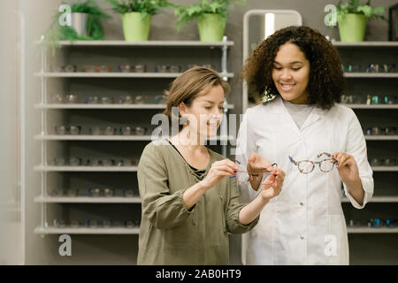 Young client of optics shop holding eyeglasses while listening to consultant Stock Photo