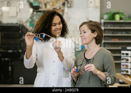 Happy young client of optics shop looking at eyeglasses held by clinician Stock Photo