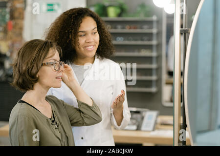 Young woman trying on new eyeglasses in front of mirror in optics shop