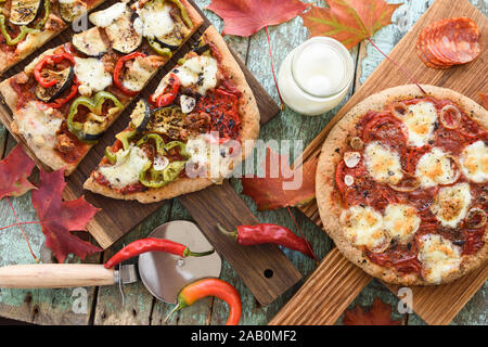 Comfort food. Homemade pizzas with eggplants, peppers, salami and mozarella on oak boards served with raw ingredients with maple leaves above view