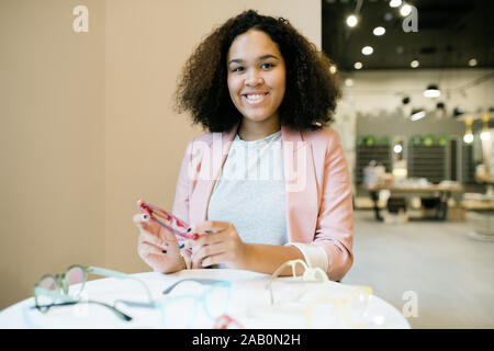 Happy young intercultural woman with eyeglasses in hands sitting in optics shop Stock Photo