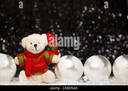 Christmas view of teddy bear in red green sweater and Christmas hat and four silver evening balls with white snow on it on dark background with silver Stock Photo