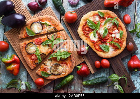 Healthy vegetarian food. Pizzas with eggplants, tomatoes and bell peppers on oak boards served with raw vegetables on shabby blue background above vie