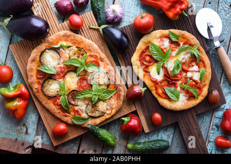 Healthy vegetarian food. Homemade Italian pizzas with aubergines, tomatoes and basil on oak cutting boards with raw vegetables on blue table above vie Stock Photo