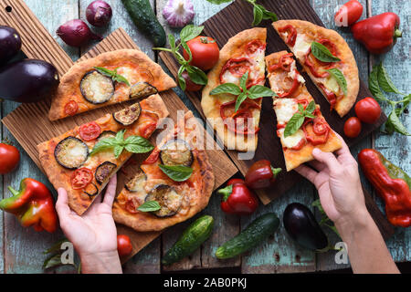 Slender woman hands reaching for healthy vegetarian pizza. Homemade rustic pizzas with aubergines, bell peppers, basil and tomatoes on oak boards with Stock Photo