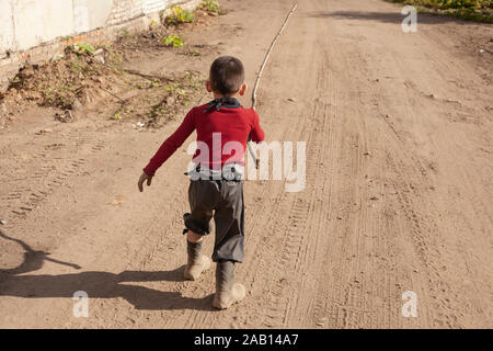 The boy runs from the pope. A child in boots and with a stick in his hands. A hyperactive child does not obey parents. The boy is dirty clothes. Child Stock Photo