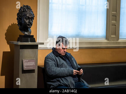 Man sitting on bench using mobile phone with sculpture of Einstein on plinth. Victoria and Albert Museum, South Kensington, London, England, UK. Stock Photo