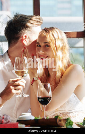 Beautiful young smiling woman drinking wine with her boyfriend and listening to him telling compliments Stock Photo