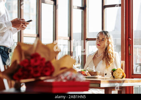 Sad pretty young woman sitting at restaurant table and looking at boyfriend texting instead of spending time with her at dinner Stock Photo