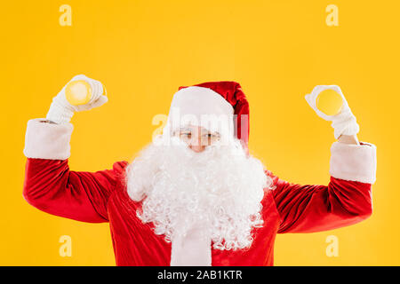 Santa Claus exercising with small dumbbells over yellow background Stock Photo