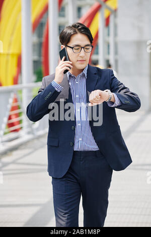 Unsmiling businessman in glasses checking time on his wristwatch when talking on phone and hurrying to meeting with business partner Stock Photo
