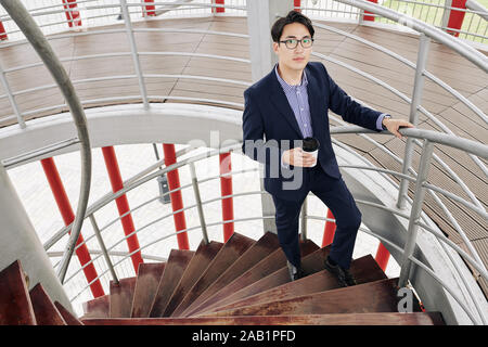 Handsome young Asian businessman with coffee cup standing on stairs in office building Stock Photo