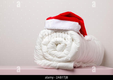 Close-up of white warm duvet roll with a cap of Santa Claus on the table against the background of light wall. Stock Photo