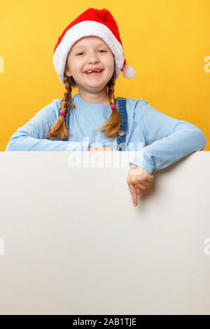 Happy child in santa hat shows his finger on an empty banner space on a yellow background. Christmas card. Stock Photo