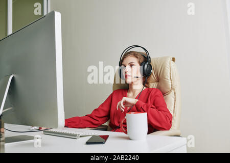 Female editor in headphones sitting at desk with big mug of coffee and working on project for video blogger Stock Photo