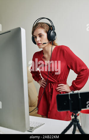 Indignant pretty young woman in red dress and headphones with microphone watching stream and making comments Stock Photo