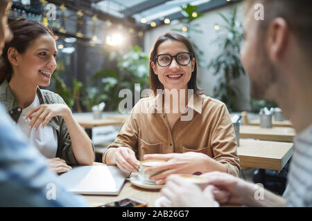 Group of contemporary young people looking at smartphone screen and laughing cheerfully while enjoying lunch together in cafe Stock Photo