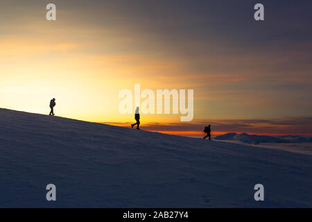 Three men descend from the mountain at sunset in winter, on a steep descent, in the distance Stock Photo