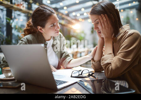 Side view portrait of frustrated young woman working at table in cafe with friend or colleague comforting her, copy space Stock Photo