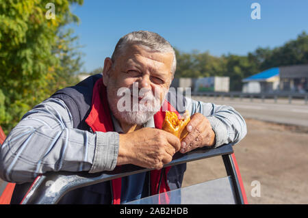 Portrait of smiling hungry senior driver eating patty leaning his elbows on the car door Stock Photo