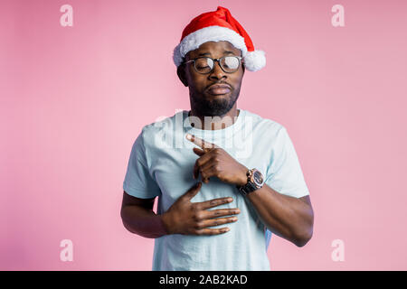 Horizontal shot of handsome bearded african male in Santa hat, dressed in casual white t shirt, pointig with index finger aside, having strangely expr Stock Photo