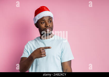 Shot of handsome serious unshaven african american young male wearing Santa hat, white t shirt, pointing with forefinger to side, standing against pin Stock Photo