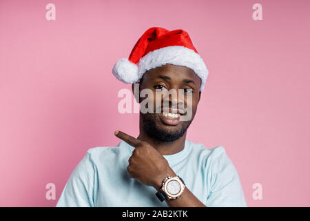 Cheerful unshaven african man wearing Santa hat, white t shirt, stylish wrist watch, pointing with index finger to side, showing something interesting Stock Photo