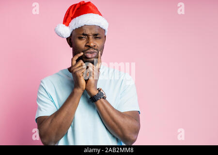 Thoughtful dark skinned man with stubble in Santa hat keeping hands on face, trying to make decision, or remember something important, thinking isolat Stock Photo
