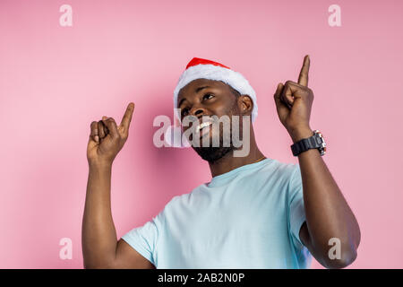 Handsome happy joyful unshaven young african american male indicating upwards with index forefingers, dressed in Santa hat and white t shirt, smiling Stock Photo