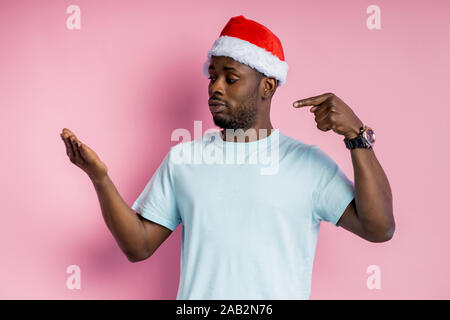 Confident attractive afro american man with stubble, wearing Santa hat, white t shirt, holding on one hand copy space and pointing it with another han Stock Photo