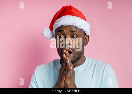 Closeup of unshaven surprised afro american man in santa hat, looks to side with shocked expression, sees something incredible, opens his mouth in ast Stock Photo