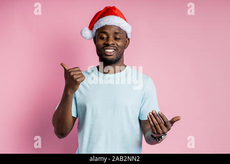 Waist up shot of satisfied unshaven african american young man in Santa hat, talking to someone, asking to see something interesting, pointing thumb t Stock Photo