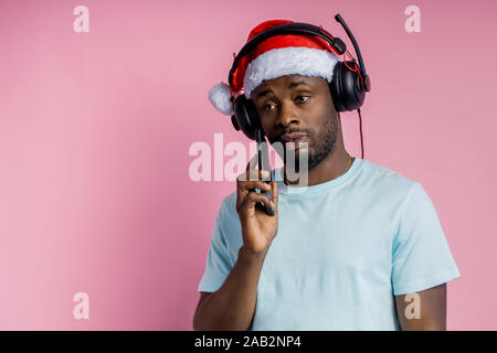 Serious sad african man wearing Santa hat, white t shirt, headphones, holding mobile phone, being in deep thought, posing on pink background with copy Stock Photo
