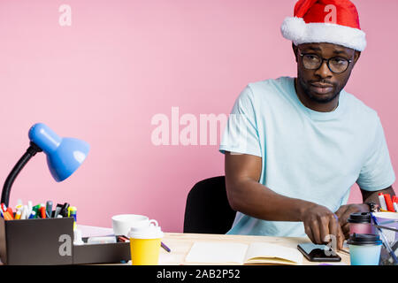 Serious young african student with small beard dressed in white t shirt, Santa Hat, sitting at table with office supplies, smartphone, doing homework, Stock Photo