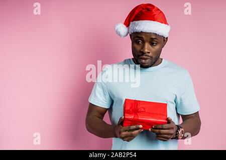 Confused puzzled handsome african american male wearing Santa hat, white t shirt, holds red box, receives unexpected gift, isolated over pink backgrou Stock Photo