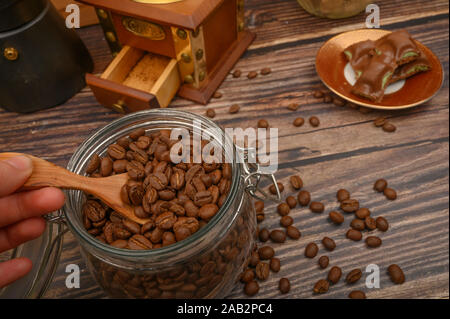 The girl's hand takes a wooden spoon of coffee beans from a glass jar, a coffee grinder, pieces of chocolate on a wooden background. Close up Stock Photo