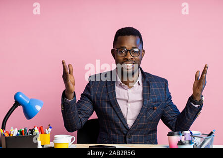 Happy cheerful young afro american employee, dressed in formal wear, sitting at desk in office, smiling, raising arms welcoming someone. People, busin Stock Photo