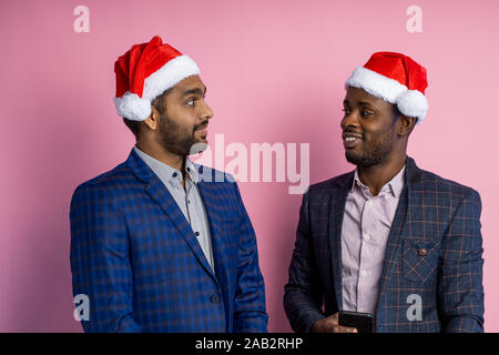 Shot of two multiethnic funny cheerful businessmen wearing suits, Santa hats, smiling, looking at each other, standing isolated over pink background. Stock Photo
