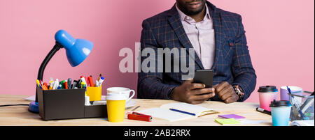 Stylish handsome afro american businessman sitting at desk in office, holding smartphone, checking email, using mobile banking, over pink background. Stock Photo