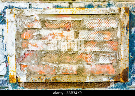 On old brick wall is insert of other bricks in rusty metal frame with peeling paint. Wall texture of orange bricks with stains of cement and stucco Stock Photo