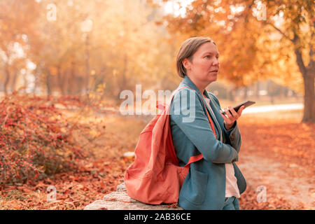 Businesswoman talking on mobile phone in autumn park, holding smartphone in front of her face, selective focus Stock Photo
