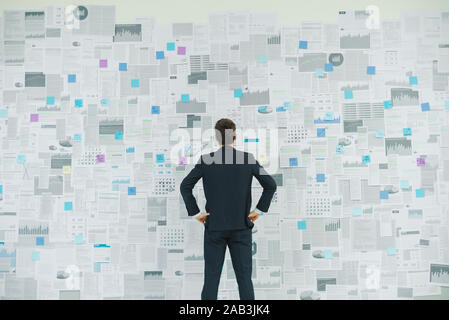 Corporate businessman standing with arms akimbo in front of a wall with lots of financial reports, business strategies and planning concept Stock Photo