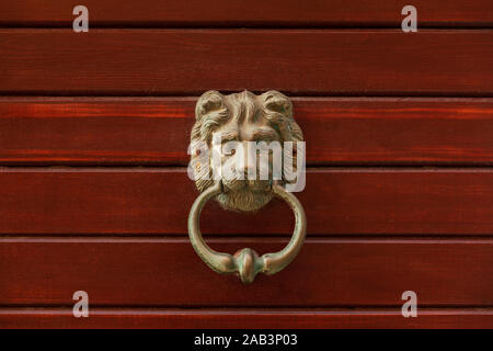 A golden brass door knob with the head of a lion holding a ring in his mouth, round handle. A wooden textured background, red colored horizontal Stock Photo